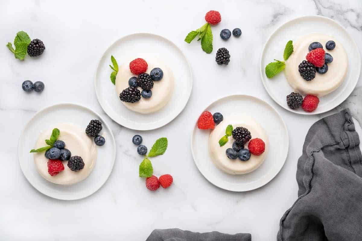 overhead view of four panna cotta on white plates with berries and mint leaves.
