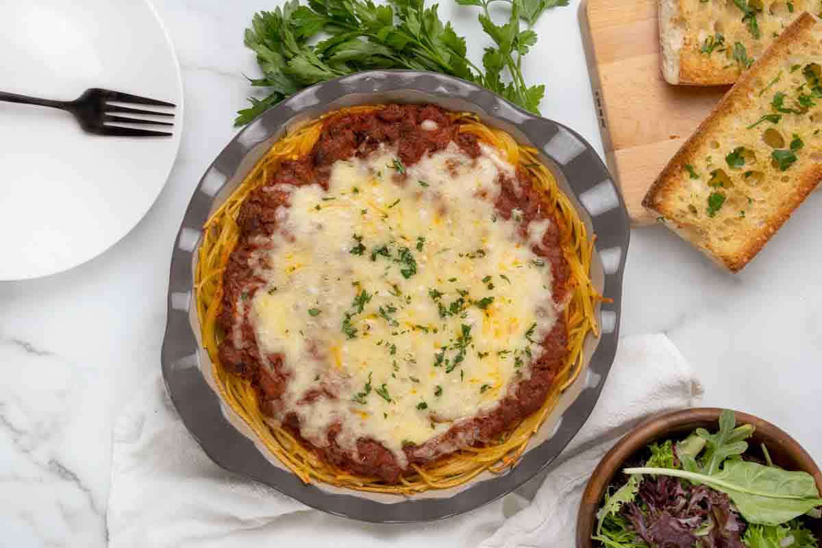whole spaghetti pie in pie plate with garlic bread and salad on the side.