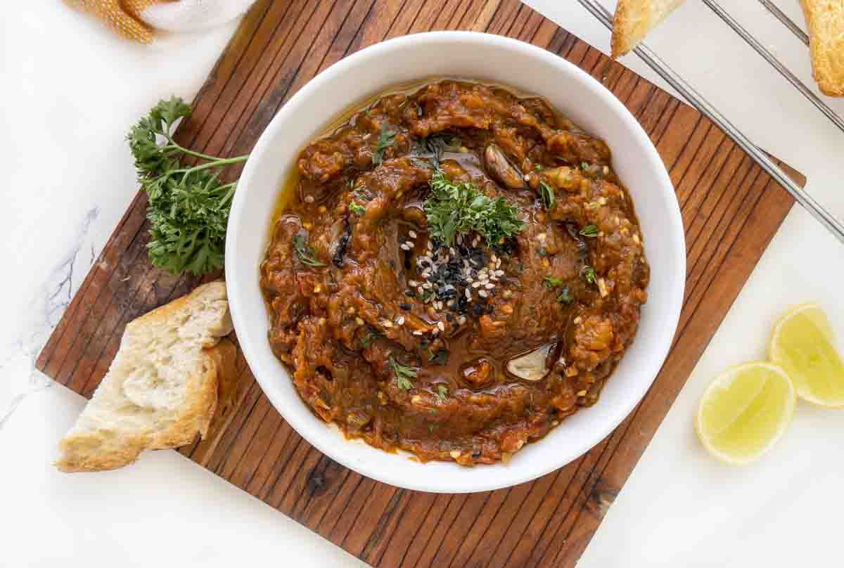 roasted eggplant dip in a white bowl with lemons and bread around it.