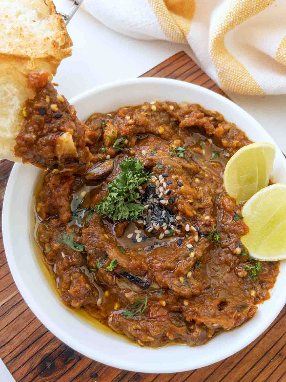roasted eggplant dip in a white bowl with lemons and a piece of bread dipping into the mixture.