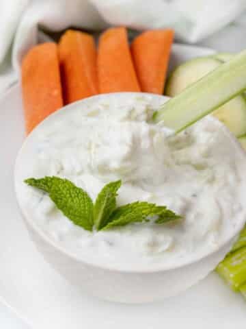 tzatziki sauce in a white bowl with a sprig of mint and carrots, and celery on a white plate.