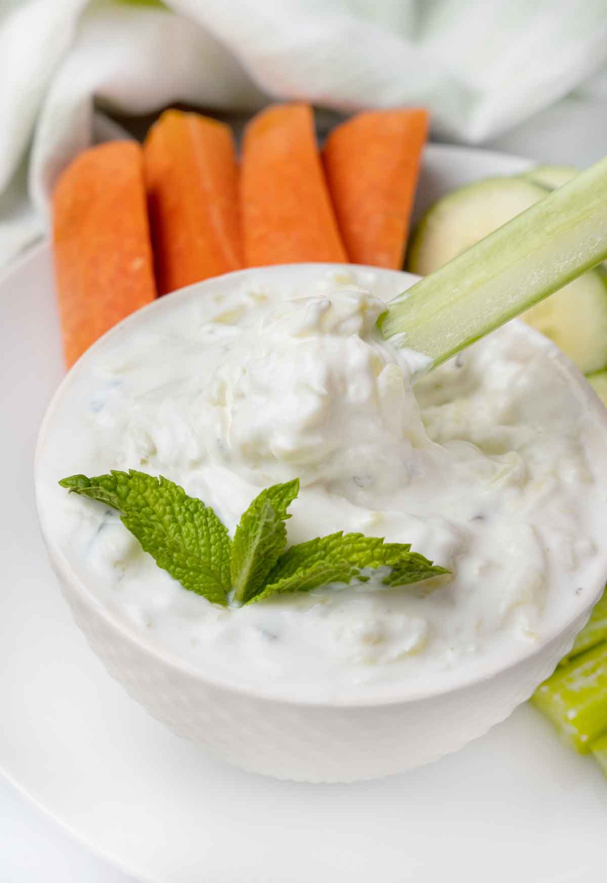 tzatziki sauce in a white bowl with a sprig of mint and carrots, and celery on a white plate.