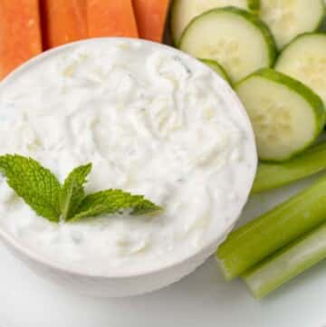 tzatziki sauce in a white bowl with a sprig of mint and carrots, cucumbers and celery on a white plate.