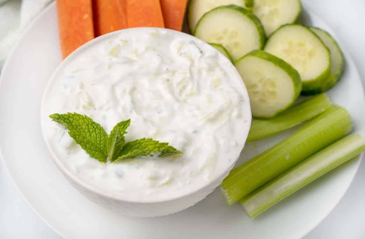 tzatziki sauce in a white bowl with a sprig of mint and carrots, cucumbers and celery on a white plate.