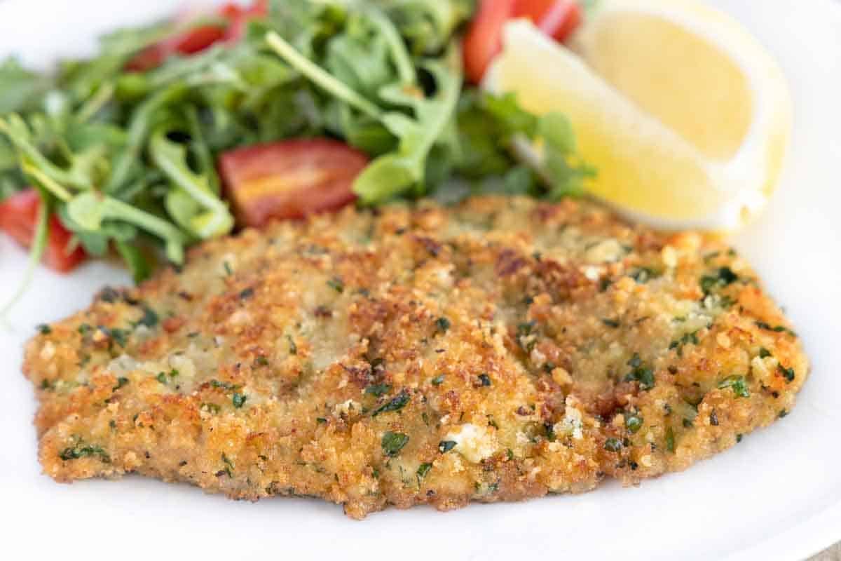 Veal Milanese on white plate with salad and lemon wedges.