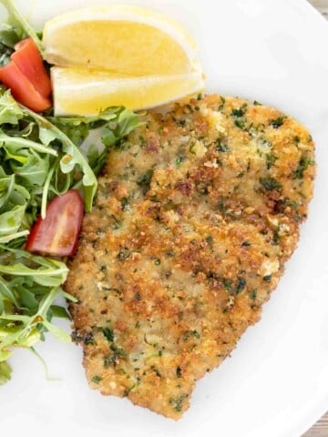 Veal Milanese on white plate with salad and lemon wedges.