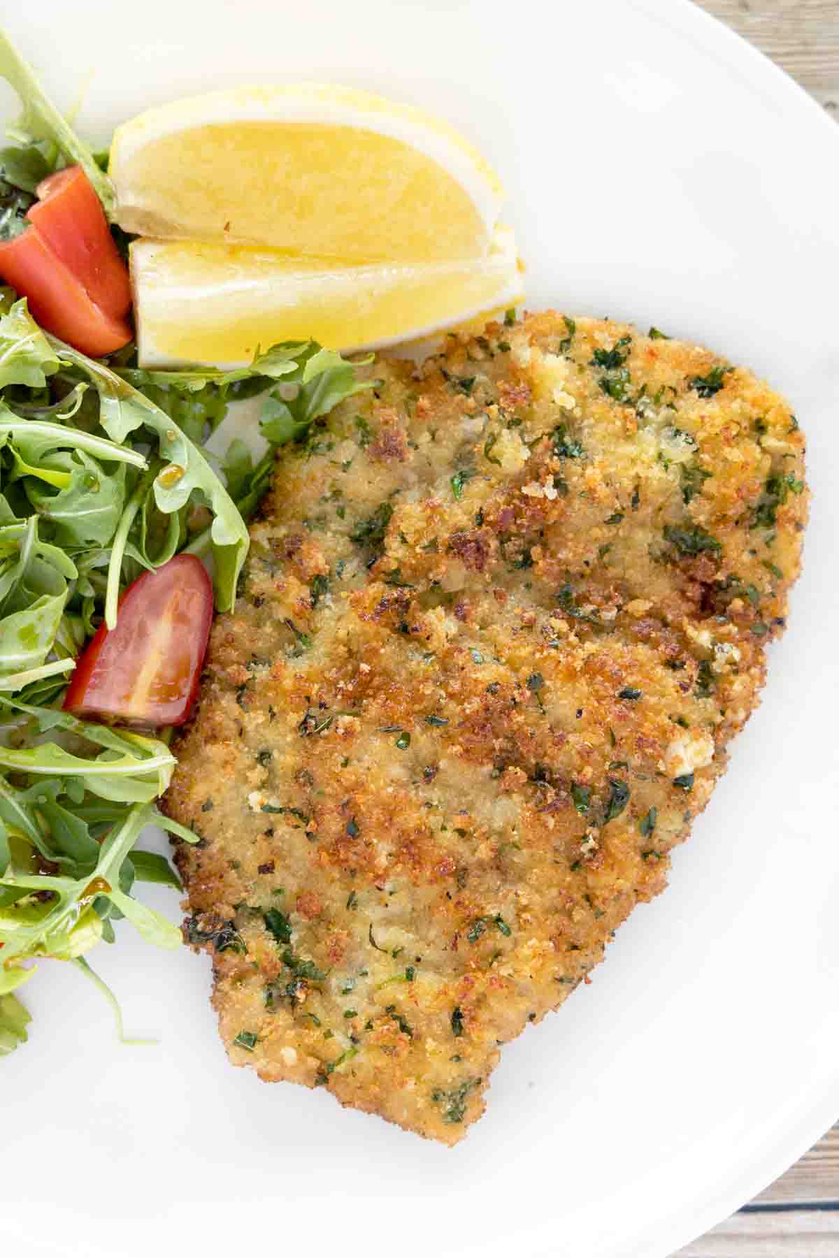 Veal Milanese on white plate with salad and lemon wedges.