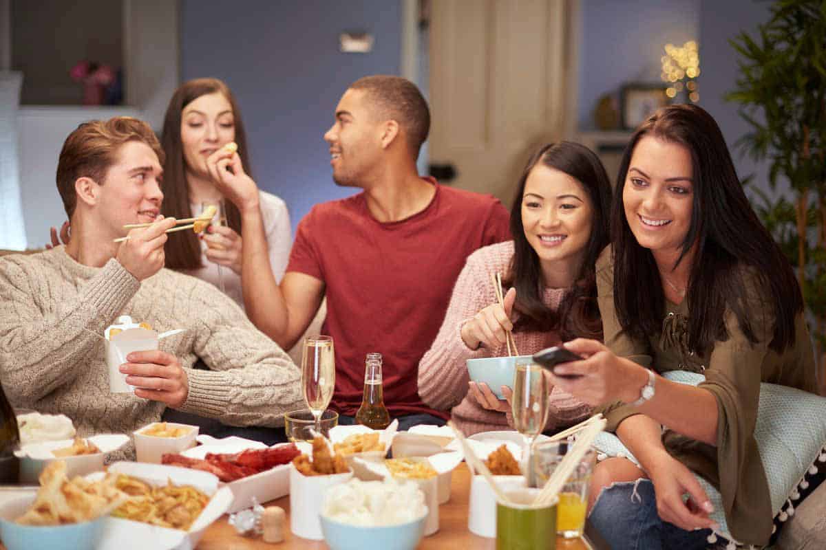 a group of people sitting around a coffee table eating as they watch tv.