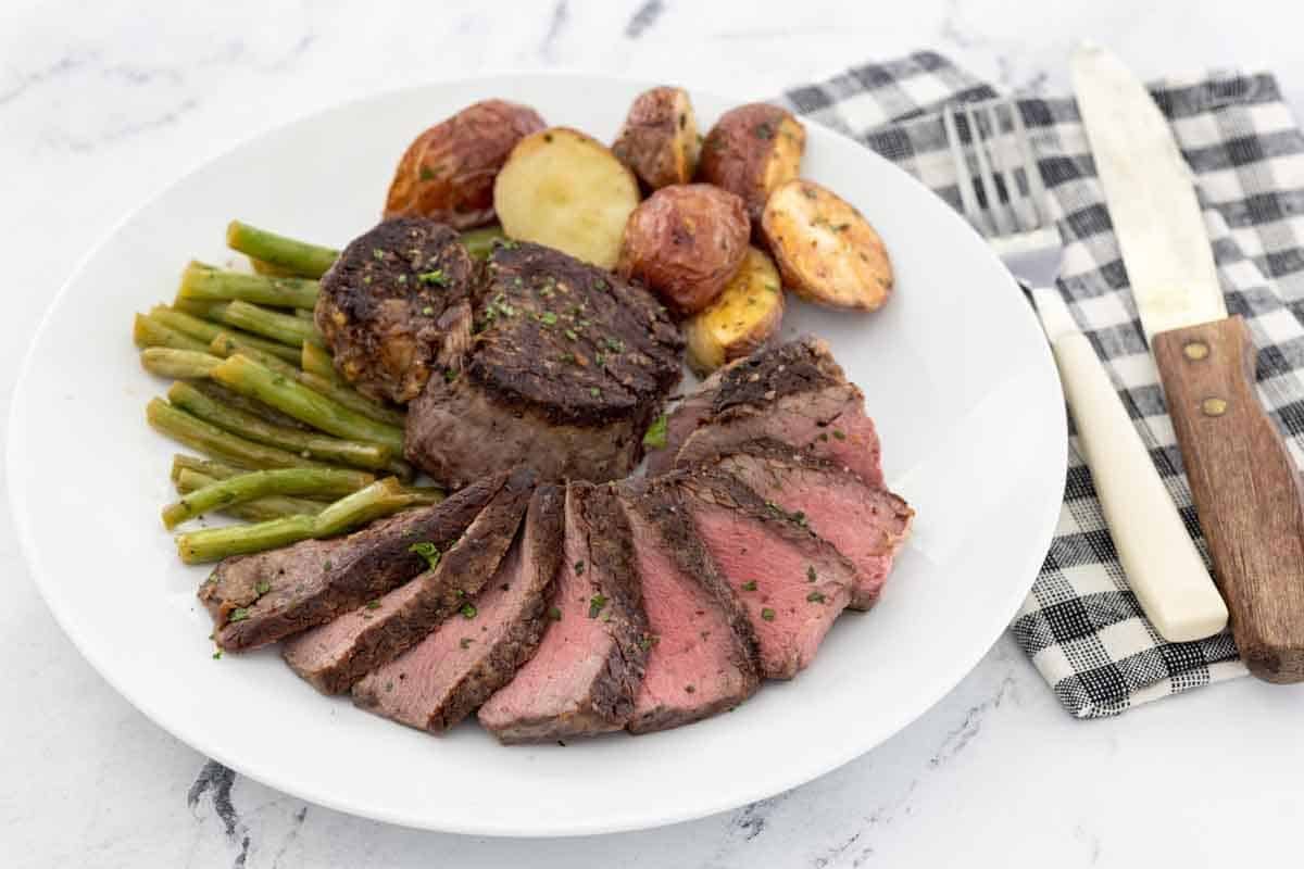 Sliced pan seared filet mignon on a white plate with potatoes and green beans.