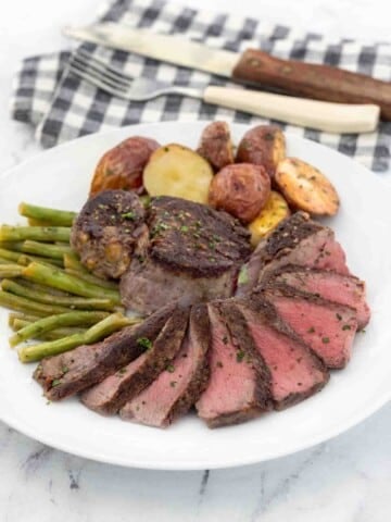 Sliced pan seared filet mignon on a white plate with potatoes and green beans.