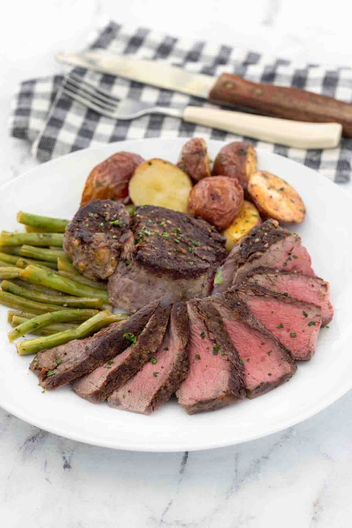 Sliced pan seared filet mignon on a white plate with potatoes and green beans.