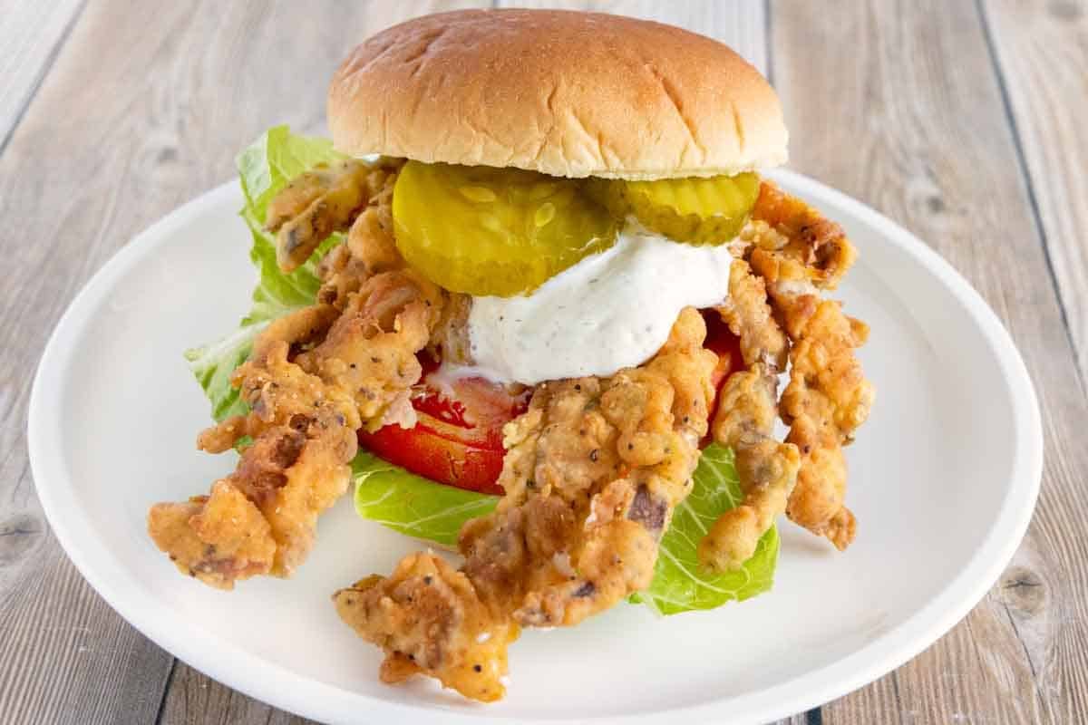 soft shell crab sandwich with lettuce, tomato, tartar sauce and pickle chips on a white plate.