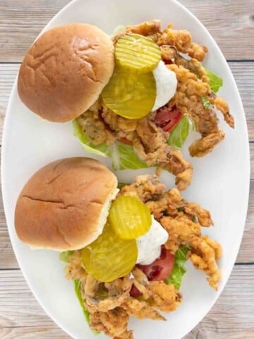 soft shell crab sandwiches with lettuce, tomato, tartar sauce and pickle chips on a white plate.