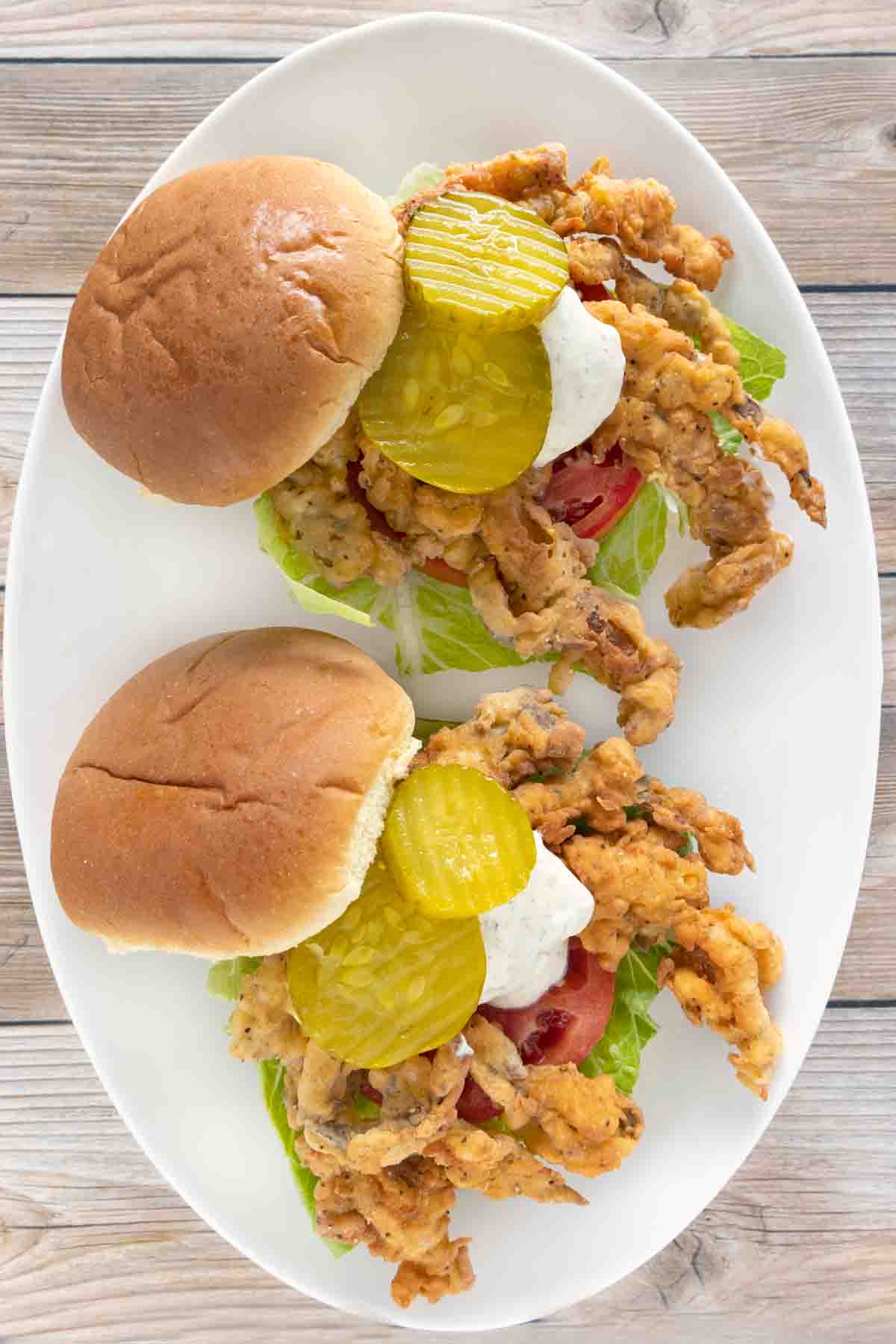 soft shell crab sandwiches with lettuce, tomato, tartar sauce and pickle chips on a white plate.