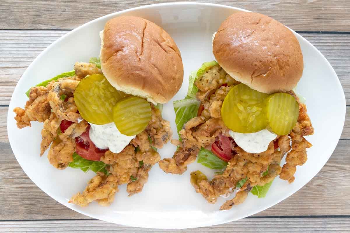 soft shell crab sandwiches with lettuce, tomato, tartar sauce and pickle chips on a white plate.