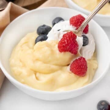 Vanilla custard with a spoon in it in a white bowl topped with whipped cream and berries.