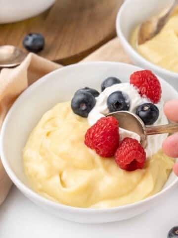 Vanilla custard with a spoon in it in a white bowl topped with whipped cream and berries.