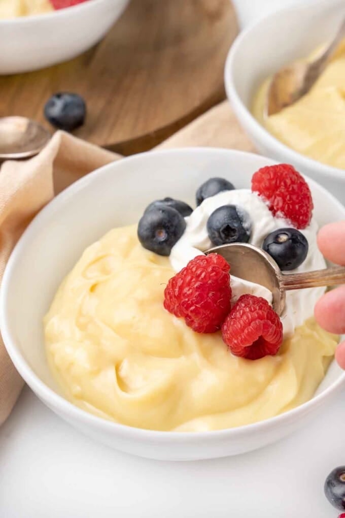 Vanilla custard with a spoon in it in a white bowl topped with whipped cream and berries.