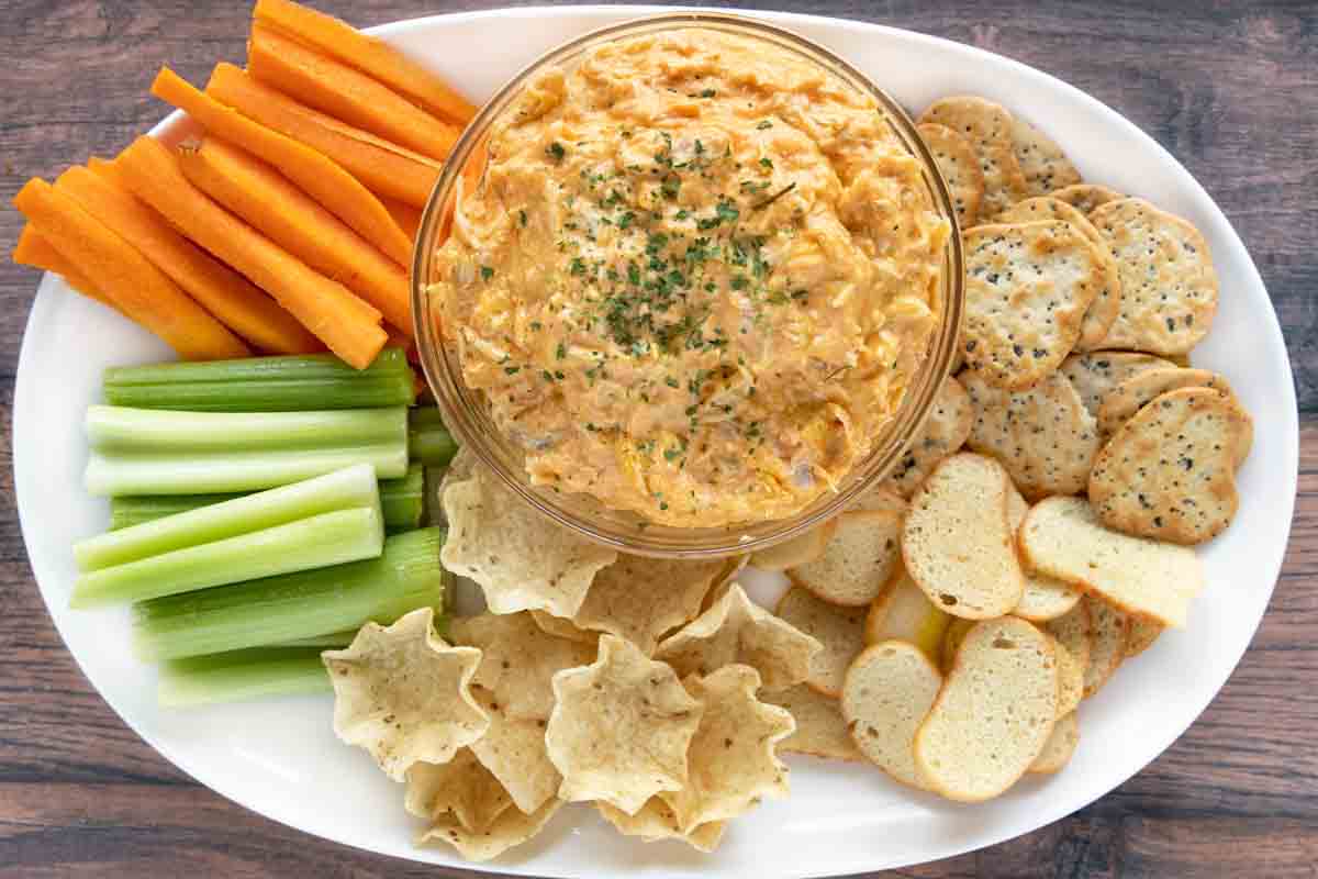 Buffalo chicken dip in a bowl on a white platter with celery , carrot sticks and crackers.