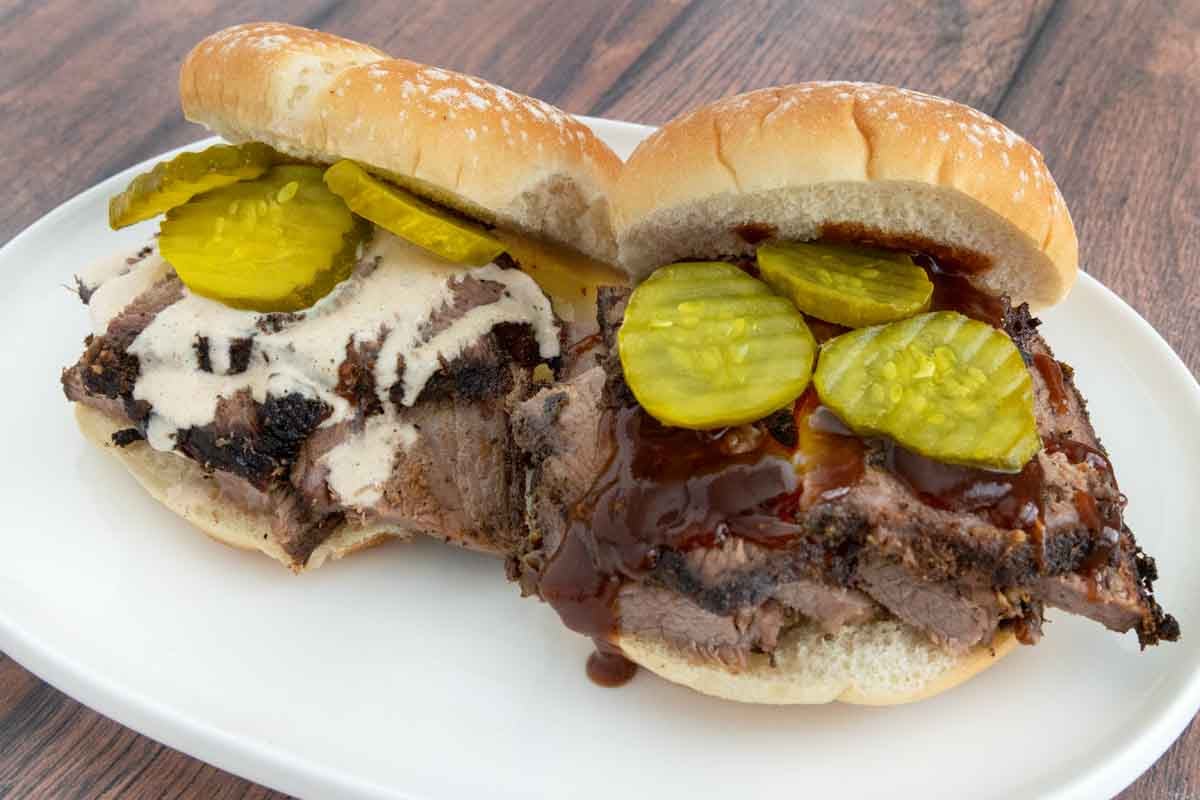 Smoked beef brisket sandwiches with bbq sauce and pickle chips on a white platter.