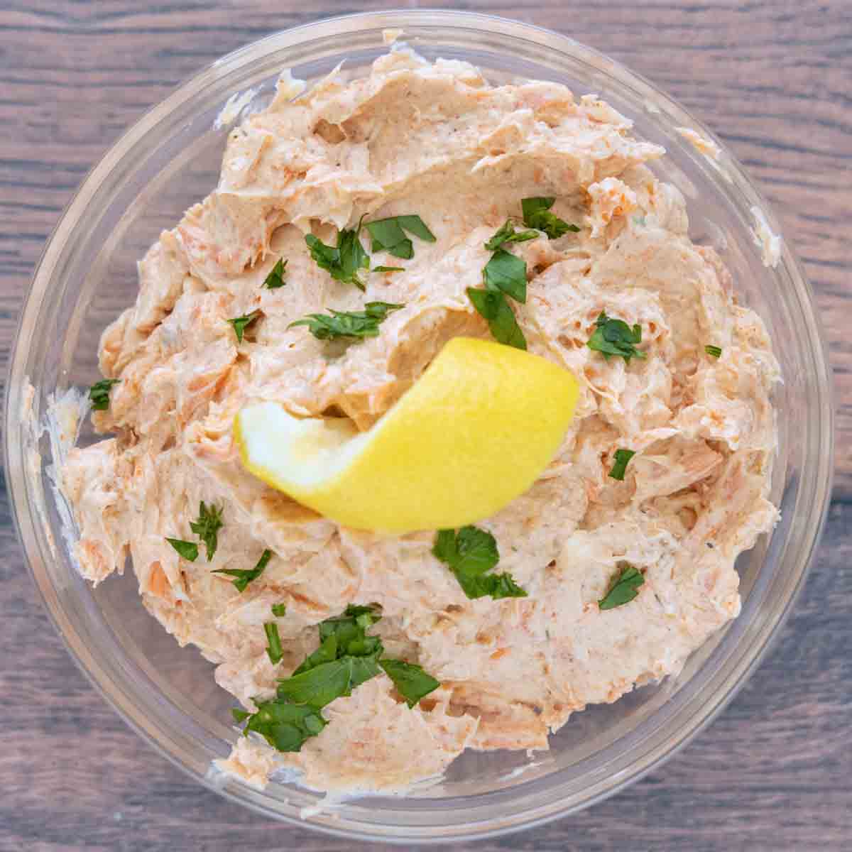 Smoked trout dip in a clear glass bowl with a lemon peel and chopped parsley.