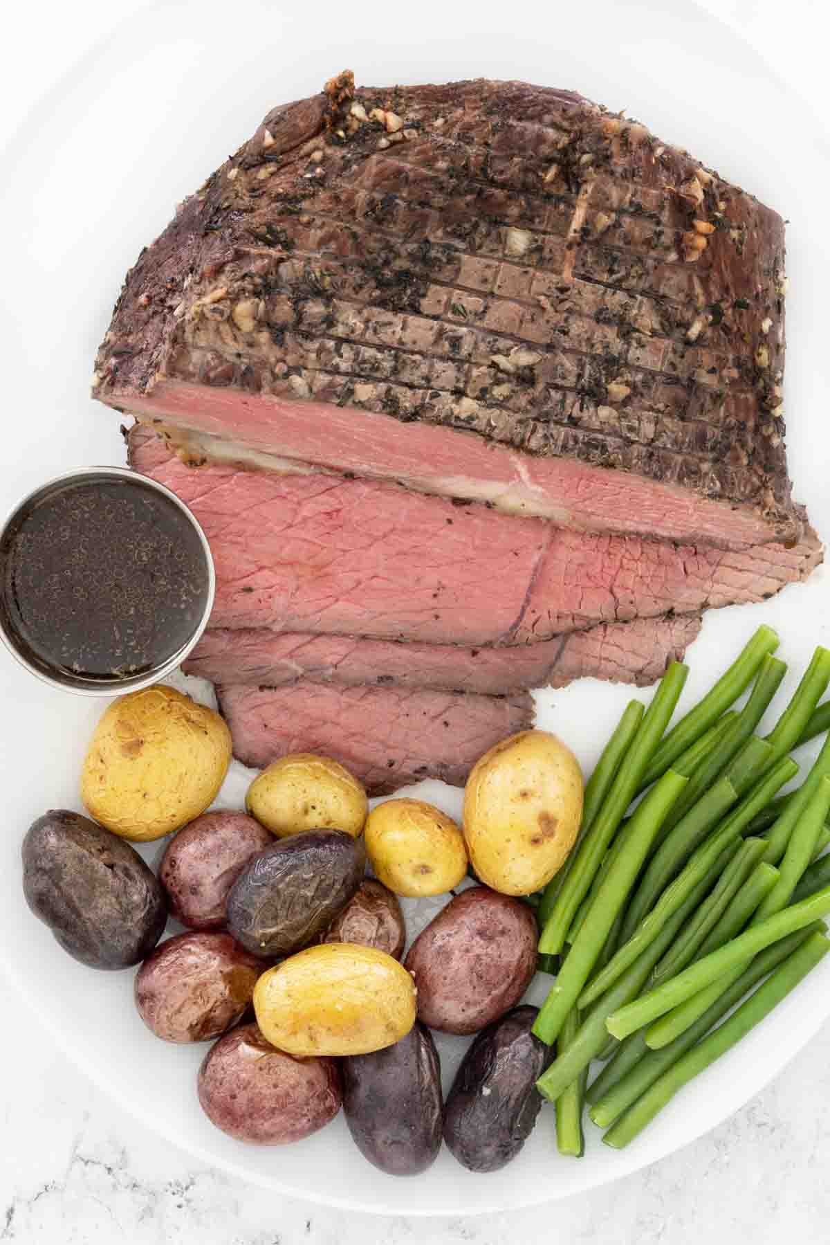 Sliced bottom round roast with potatoes and green beans on a white platter.