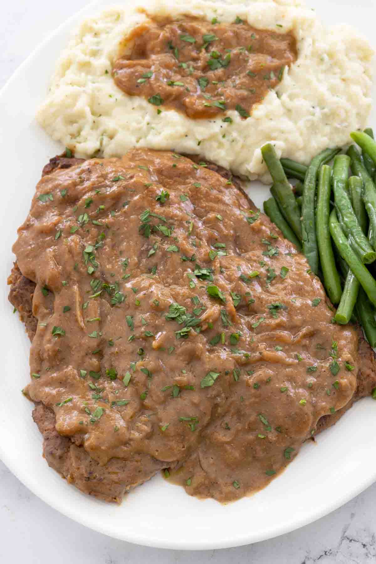 Smothered cube steak with mashed potatoes and green beans on a white plate.