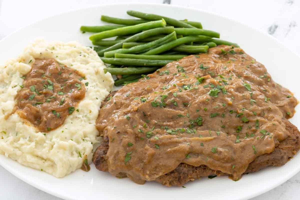 Smothered cube steak with mashed potatoes and green beans on a white plate.