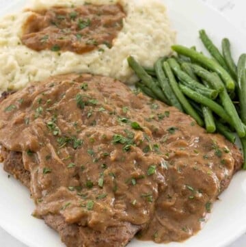 Smothered cube steak with mashed potatoes and green beans on a white plate.