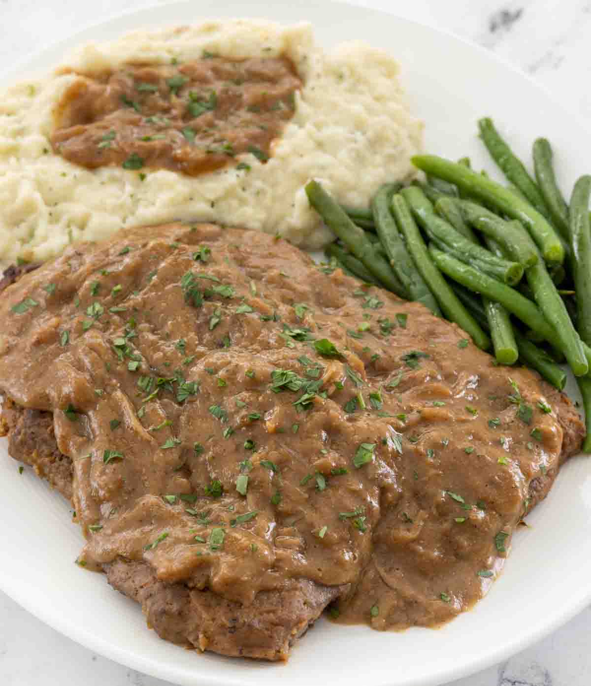 Smothered cube steak with mashed potatoes and green beans on a white plate.