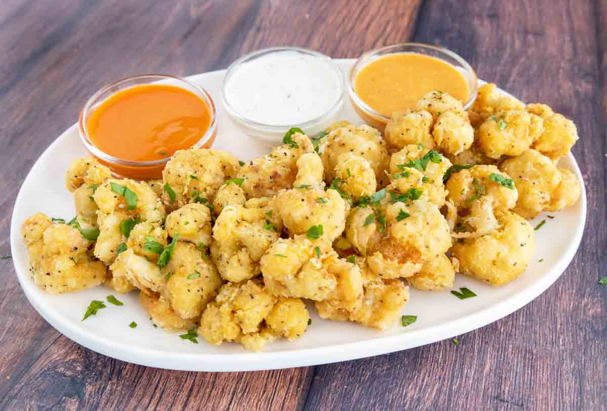 Deep fried cauliflower bites on a white platter with three dipping sauces.