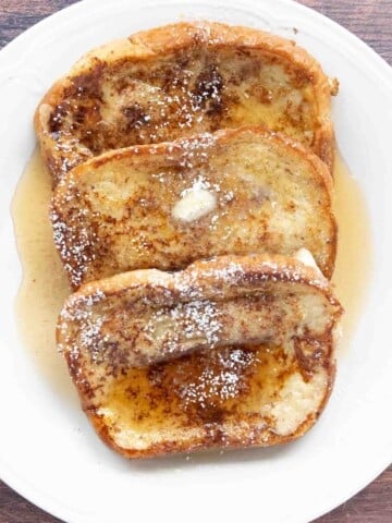 French Toast with butter, maple syrup and confectioners sugar on a white plate.