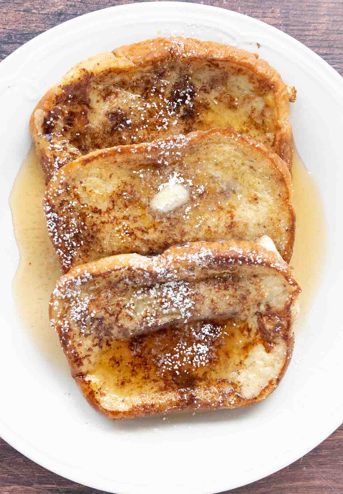 French Toast with butter, maple syrup and confectioners sugar on a white plate.