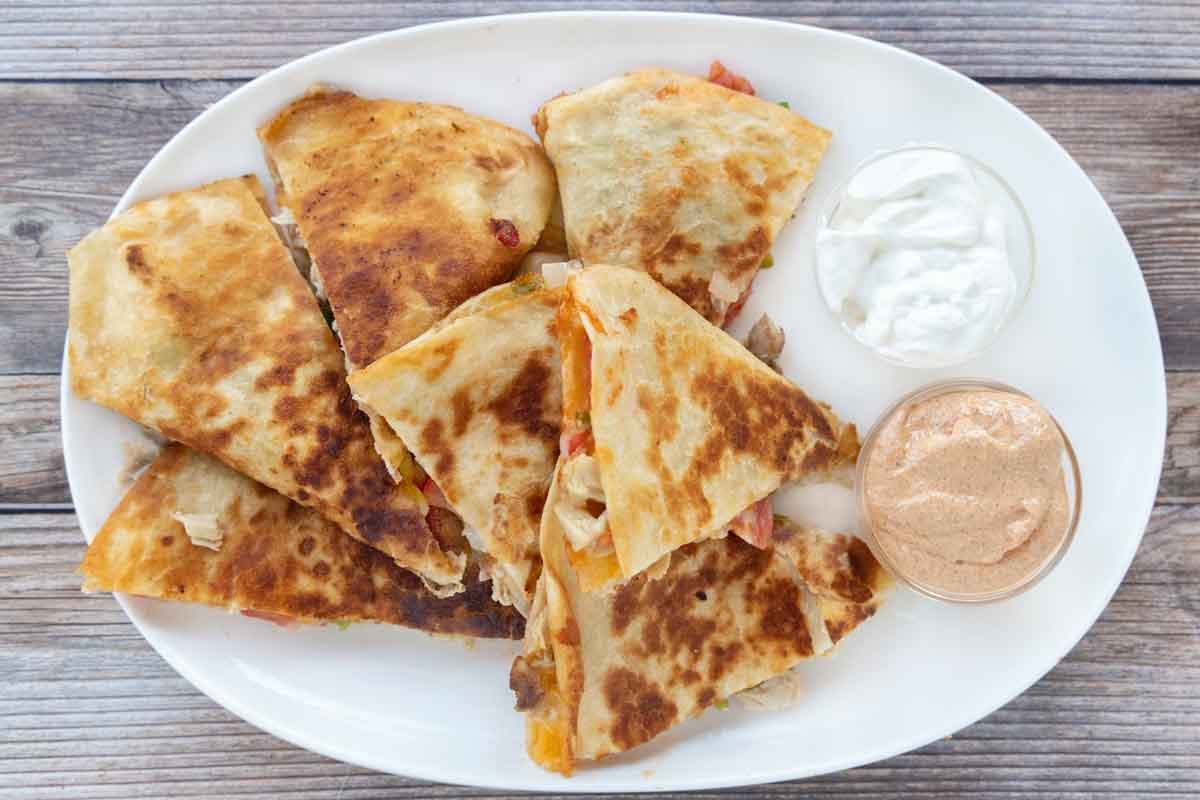 Chicken quesadilla cut into sections on a white platter with dipping sauces.