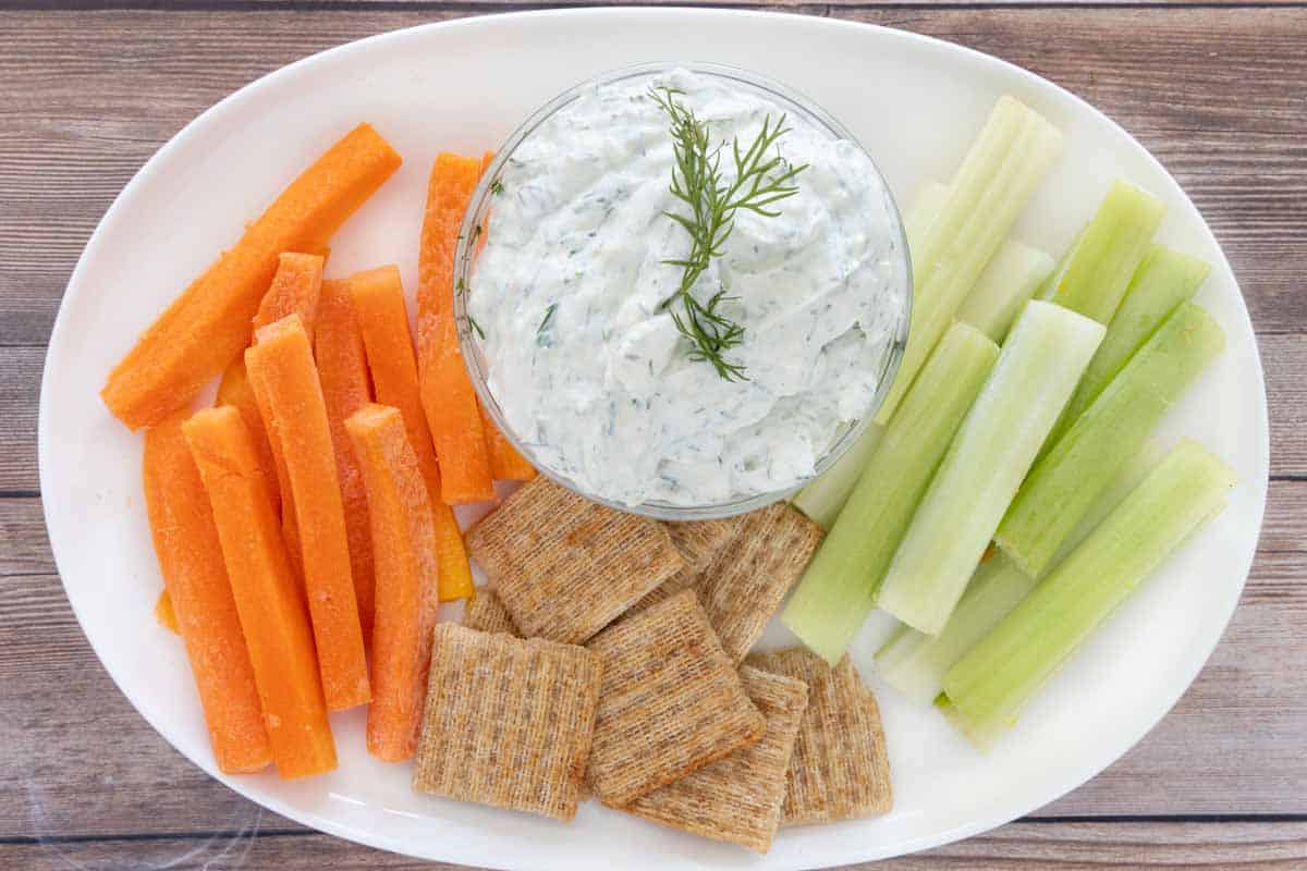 Dill yogurt sauce on a white platter with crackers, celery and carrot sticks.