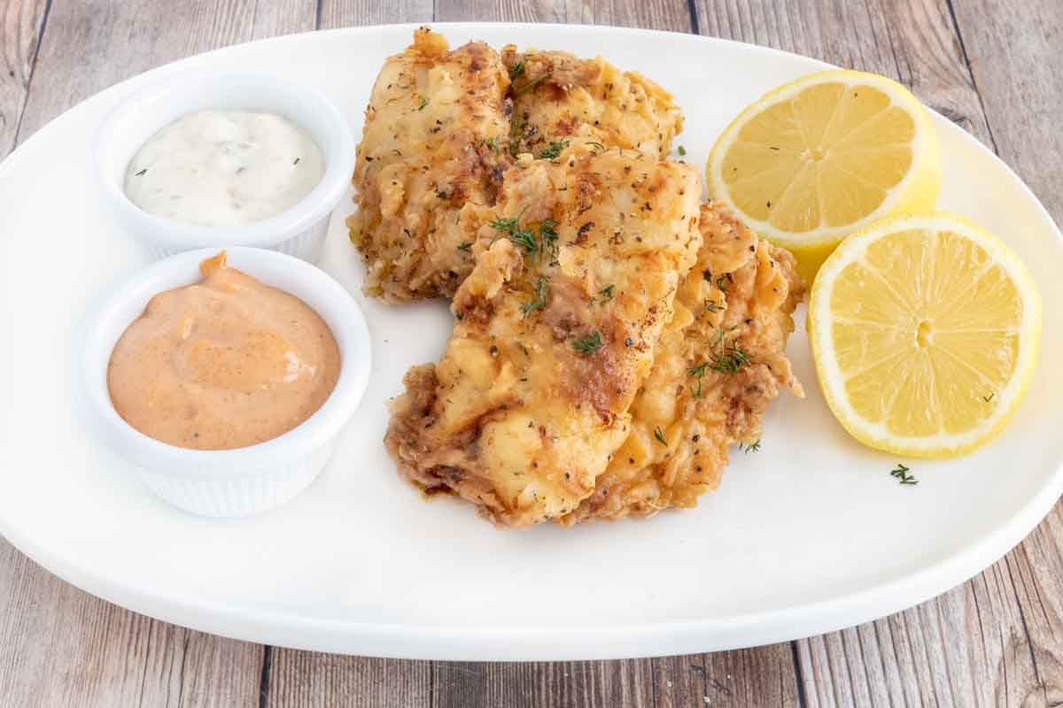 Fried walleye on a white plate with sauces and lemon halves.