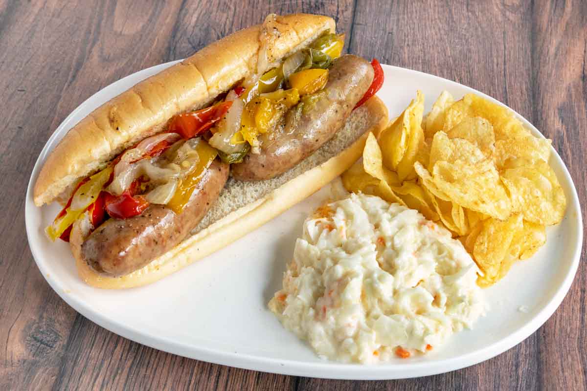 Sausage, peppers and onions hoagie,on a white plate with potato chips and coleslaw.
