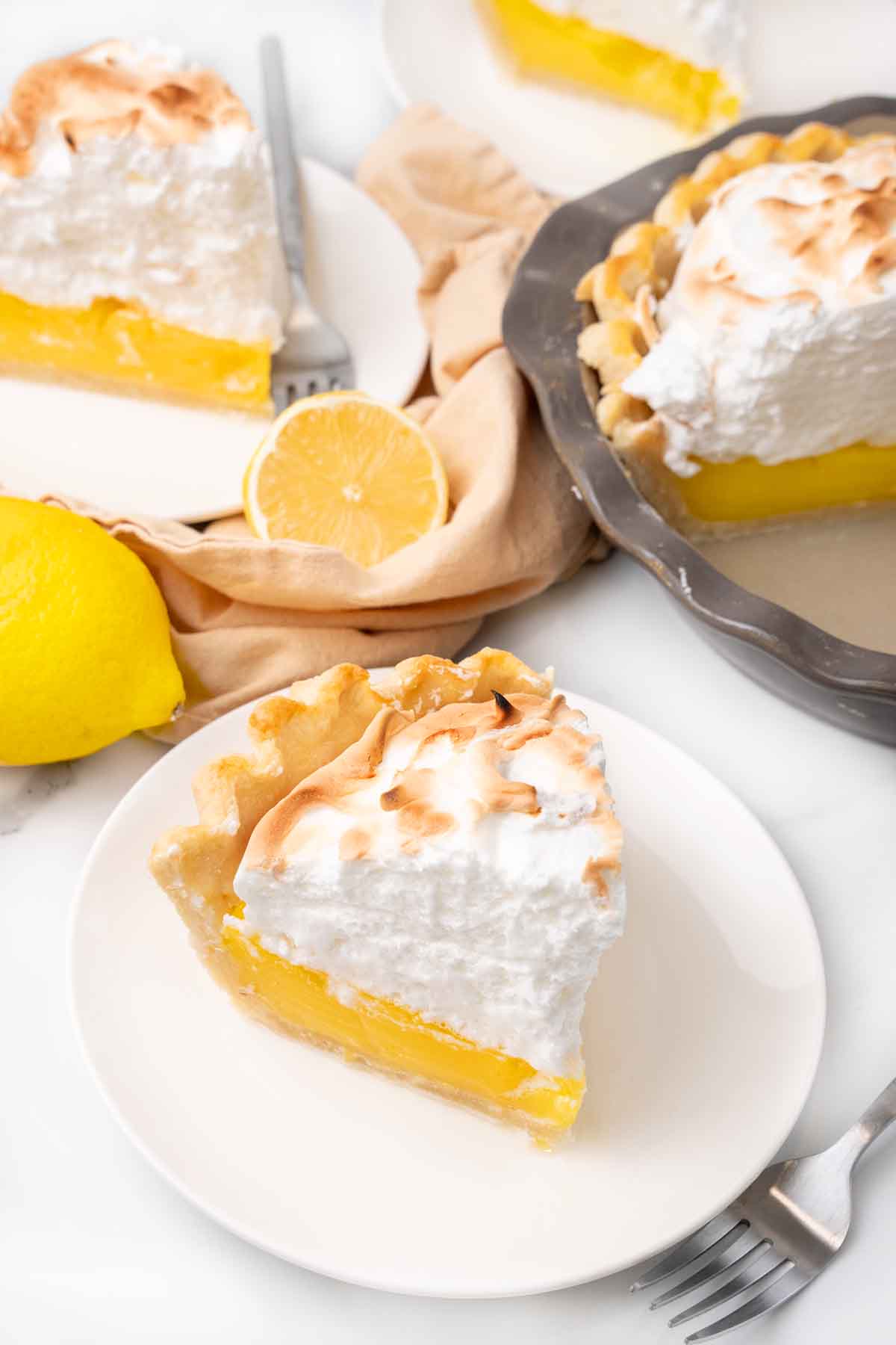 Slice of lemon meringue pie on a white plate with more pie in the background