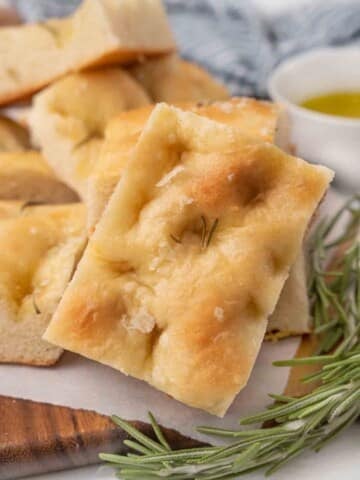 Slices of focaccia bread with sprig of rosemary on a brown cutting board.