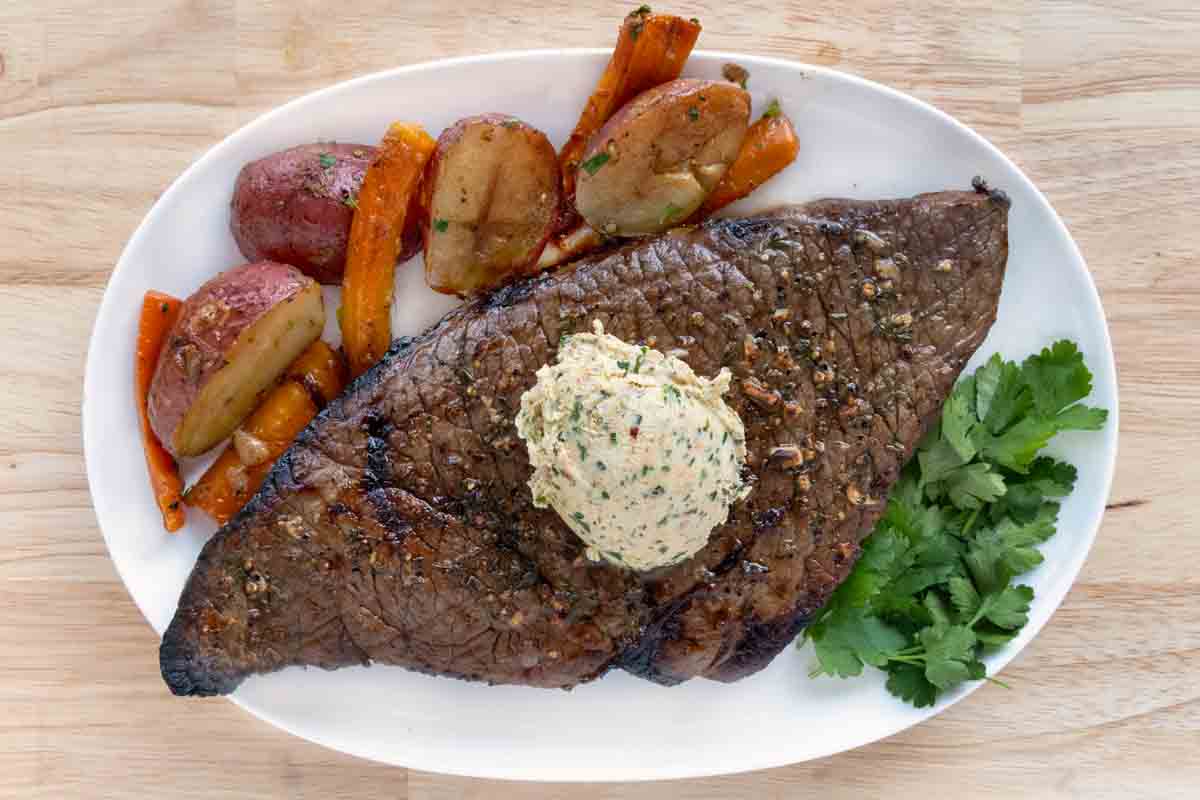 London Broil with compound butter on a white platter with roasted vegetables.