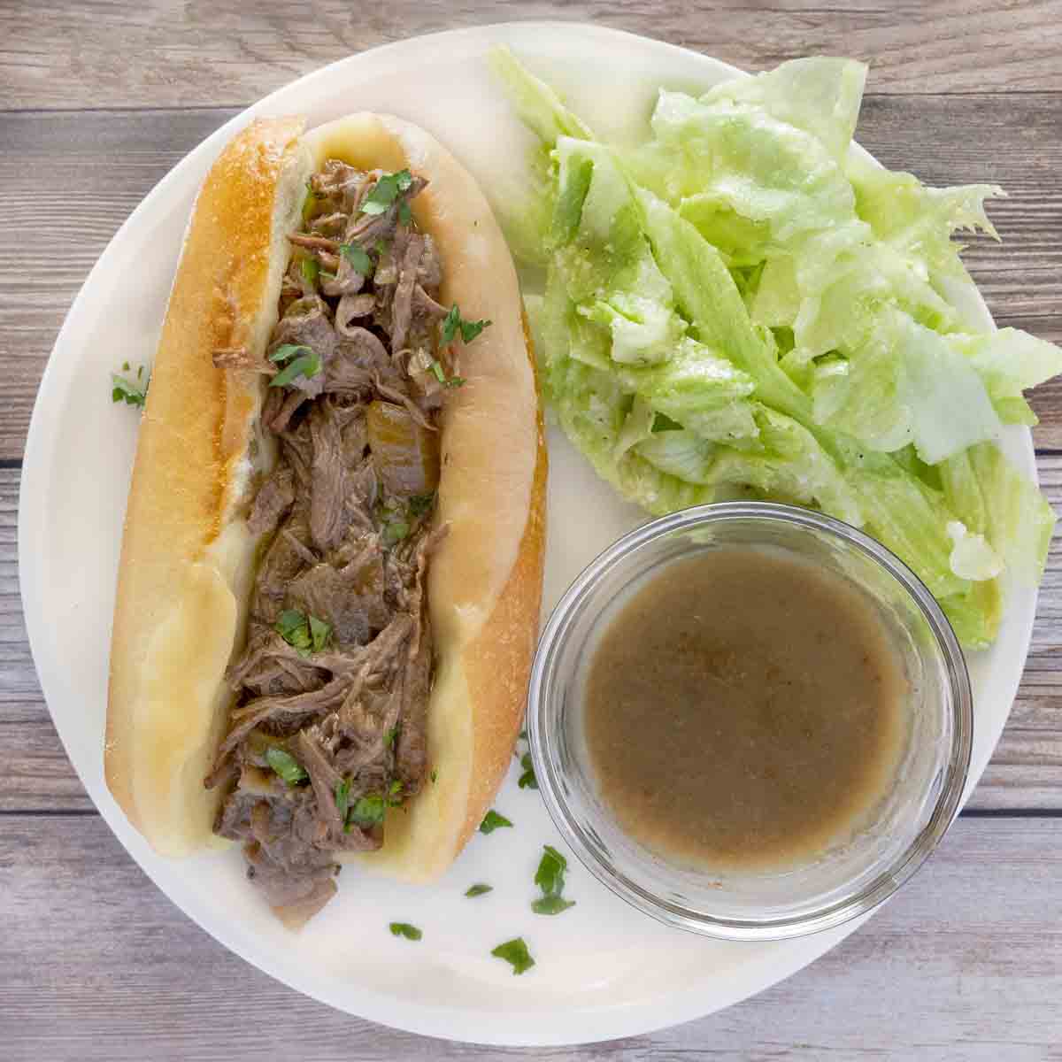 French dip sandwich on a white plate with au jus and salad.