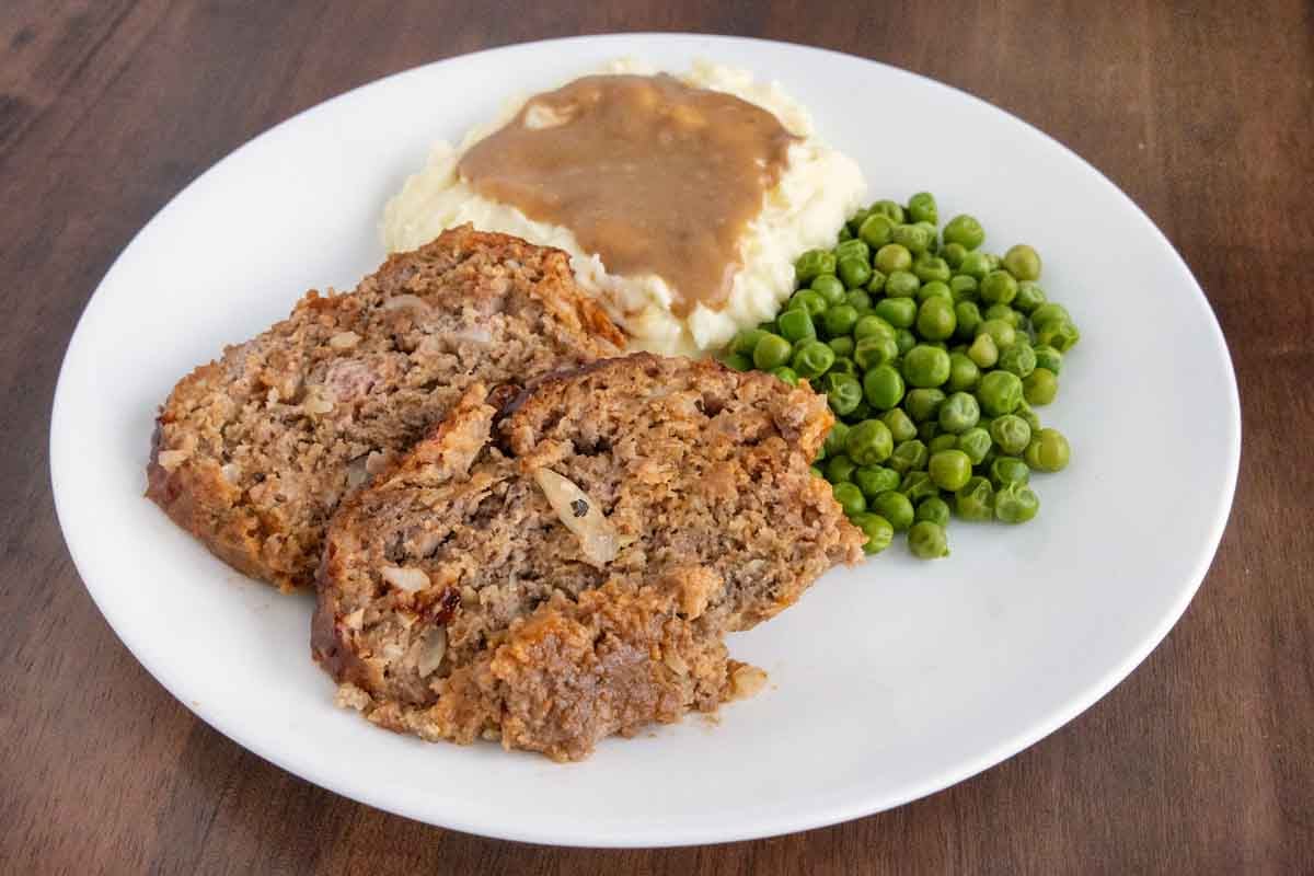 2 slices of Lipton French onion meatloaf with mashed potatoes and peas on a white plate.