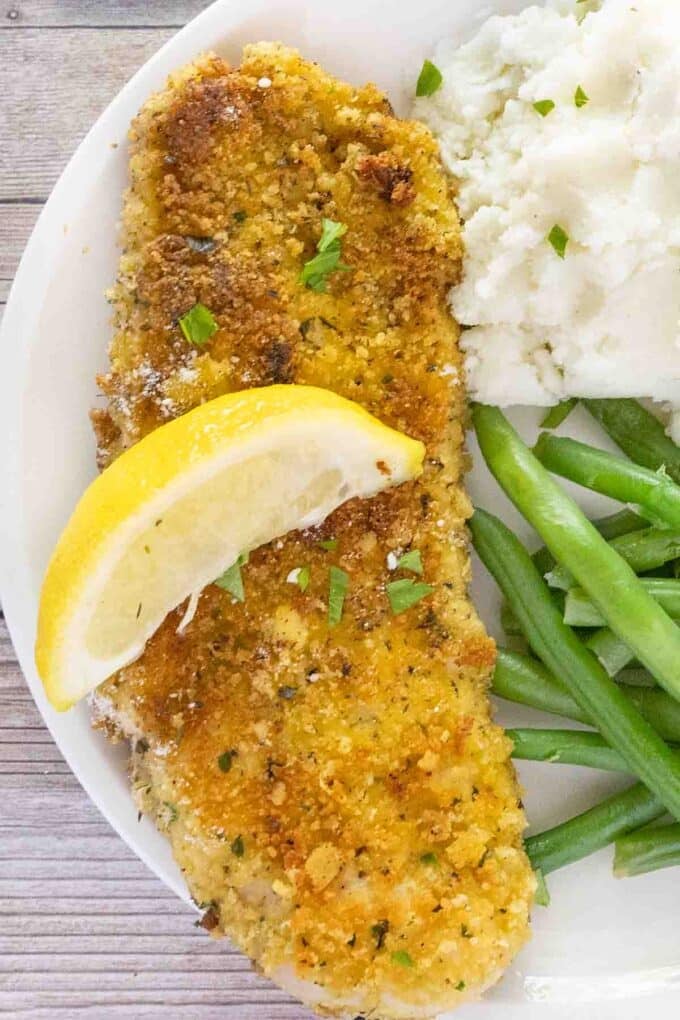 Parmesan crusted chicken with a lemon wedge on a white plate with mashed potatoes and green beans.