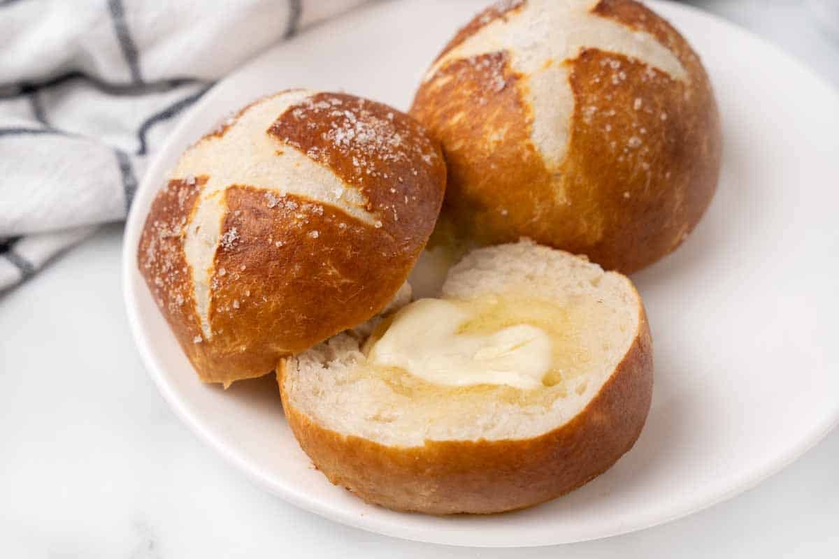 Two pretzel rolls on a white plate with one sliced with butter melting into it.