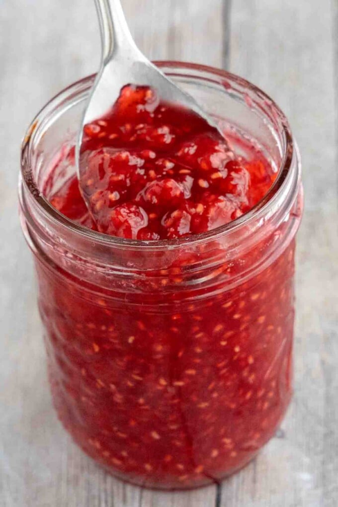 Raspberry Jam in a glass jar with a spoon coming out.