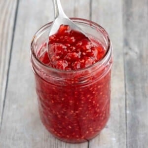 Raspberry Jam in a glass jar with a spoon coming out.