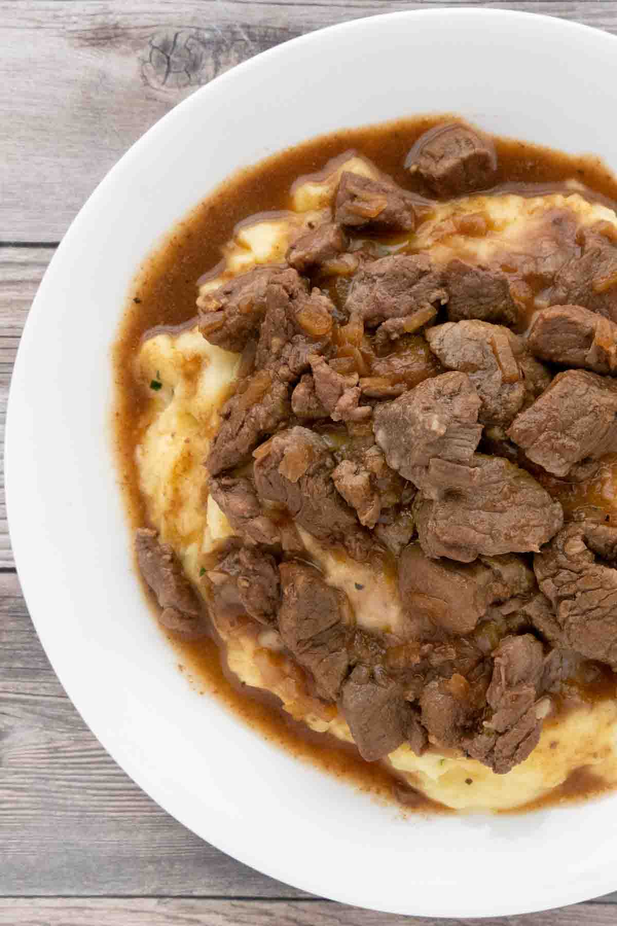 Slow Cooker Beef Tips over mashed potatoes in a white bowl.