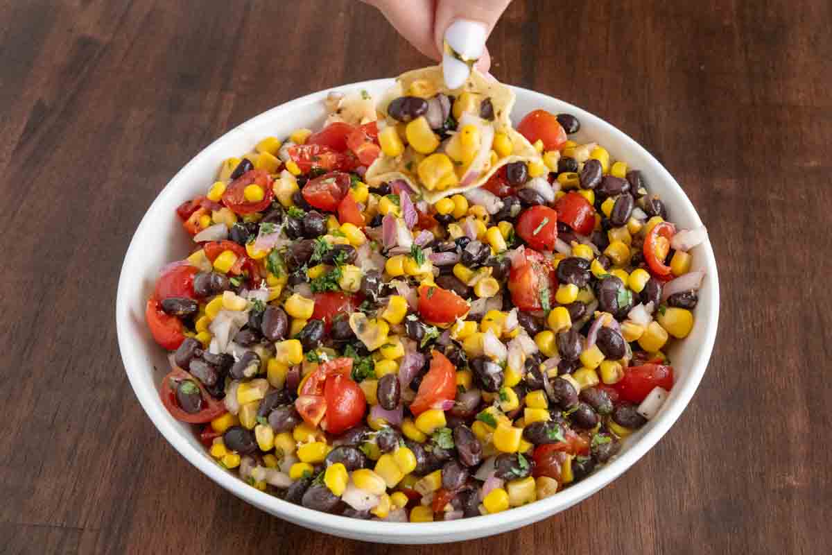 Black Bean and Corn Salsa Dip in a white bowl with a hand holdng a chip.