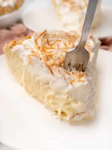 Slice of coconut cream pie with a fork in it on a white plate.