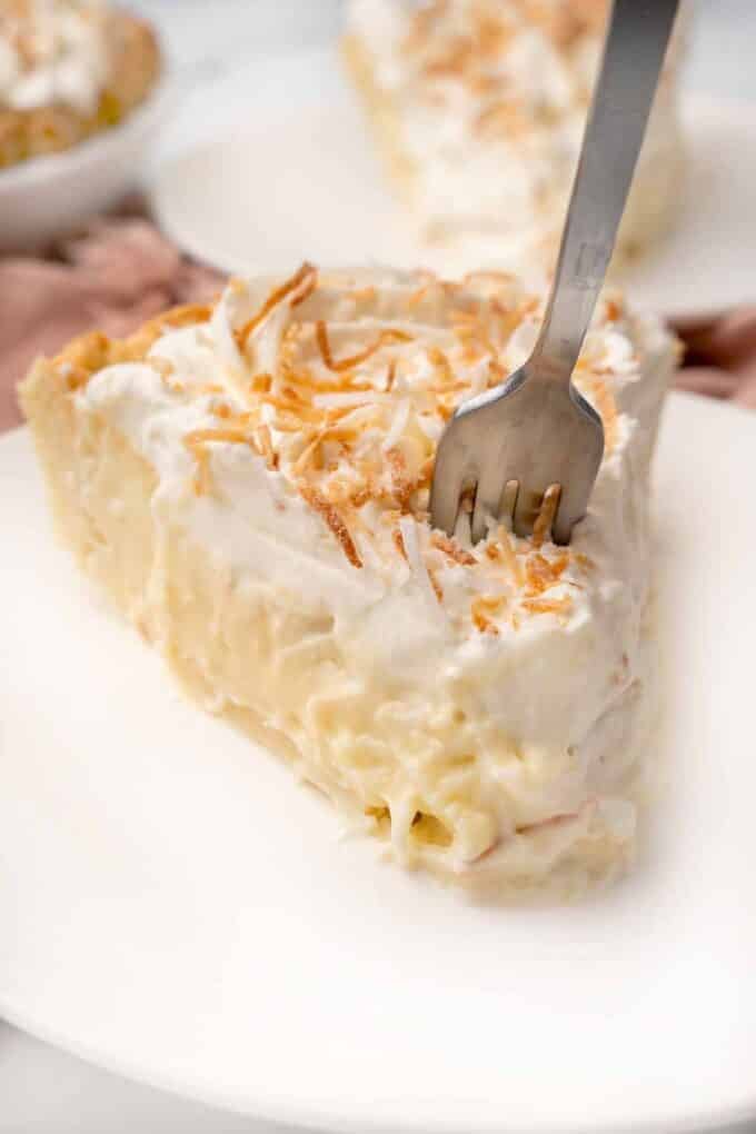 Slice of coconut cream pie with a fork in it on a white plate.